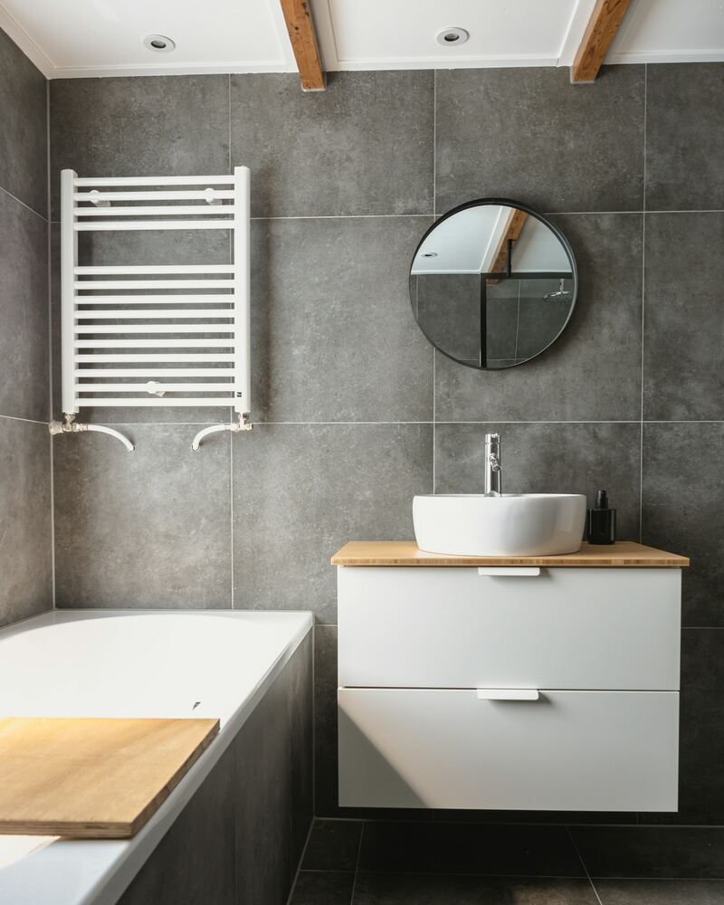 Salle de bain carrelée de gris, avec sèche-serviettes, meuble lavabo et baignoire.